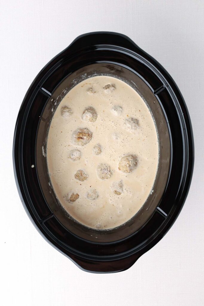 Top down image showing the inside of a slow cooker with a light almost white sacue and meatballs