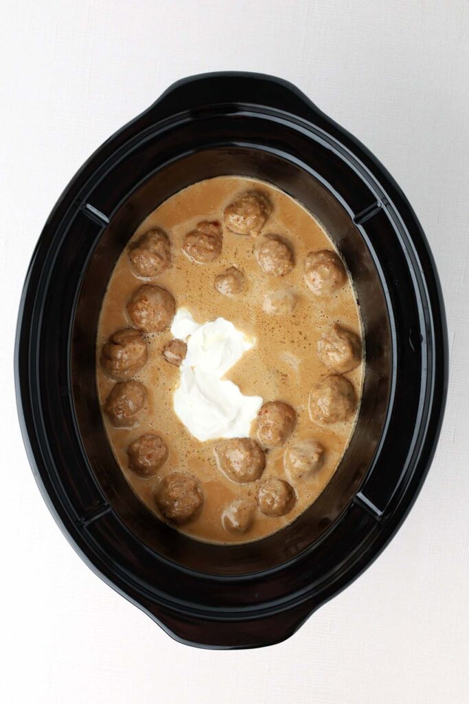 Topdown image showing the inside of a slow cooker with dark brown gravy, meatballs, and a blob of sour cream