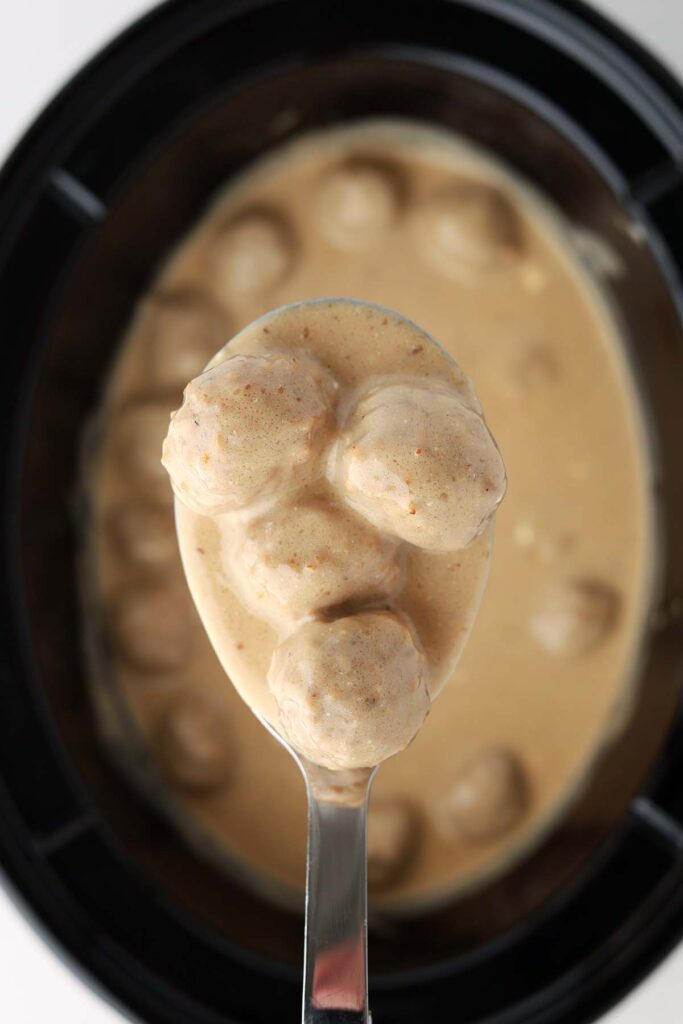 Top down image showing a serving spoon with light brown gravy and meatballs over a basin from a slow cooker
