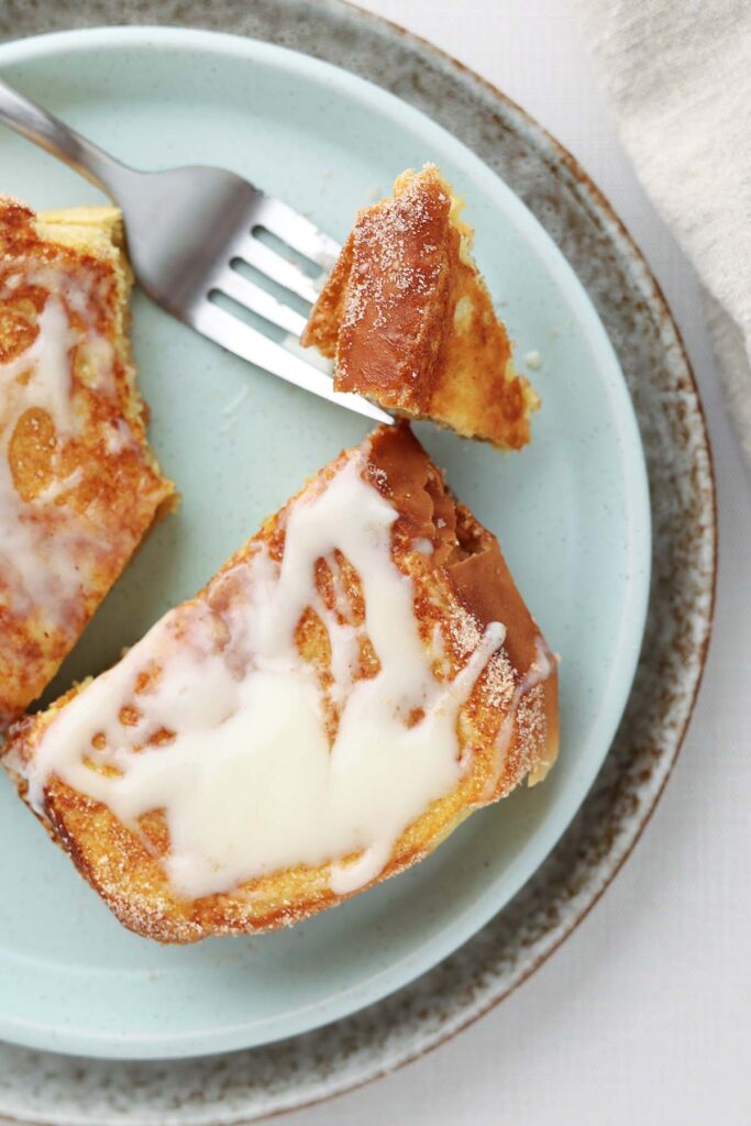 Top down image zoomed close to a french toast that has cinnamon and sugar plus a glaze. The french toast is sitting on a light blue plate. a bite on a fork