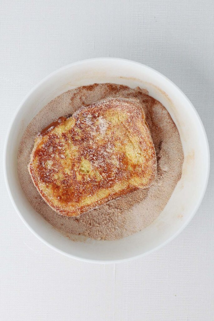 Top down image showing a golden brown french toast in a small white bowl filled with cinnamon and sugar