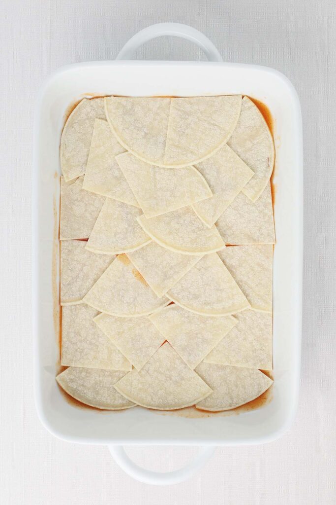 Top down image showing a white baking pan filled with enchilada sauce and a layer of cut up tortillas