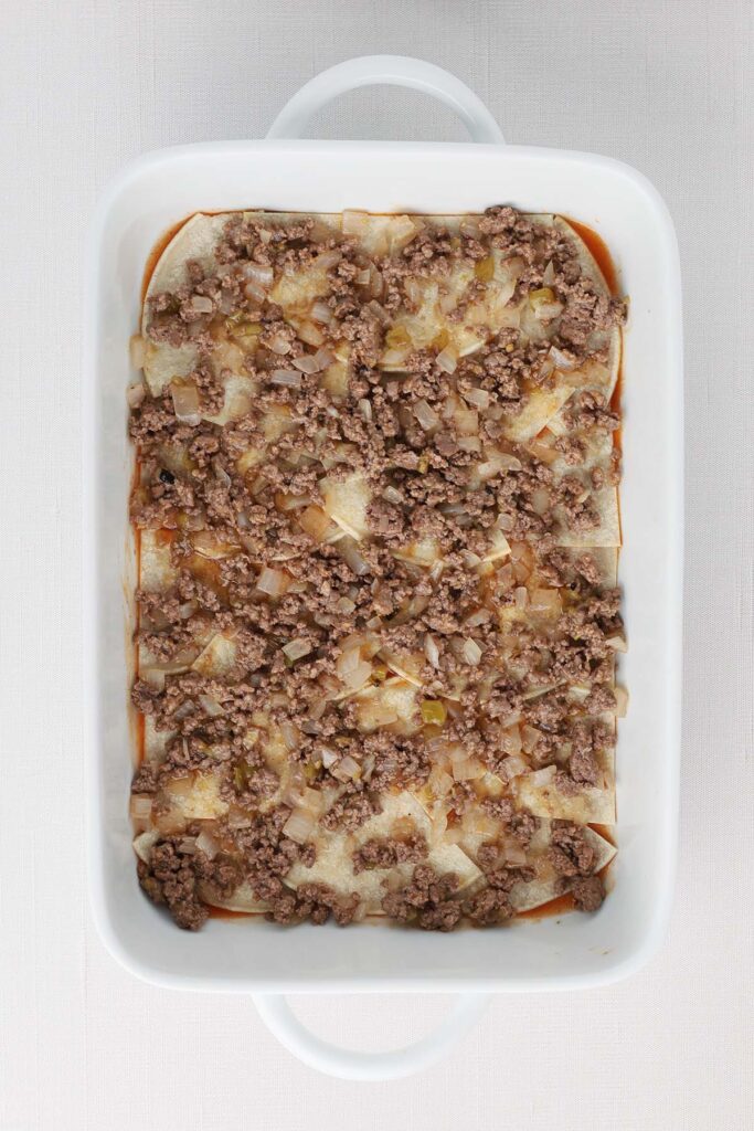 Top down image showing a white baking pan filled with enchilada sauce and a layer of cut up tortillas and ground beef