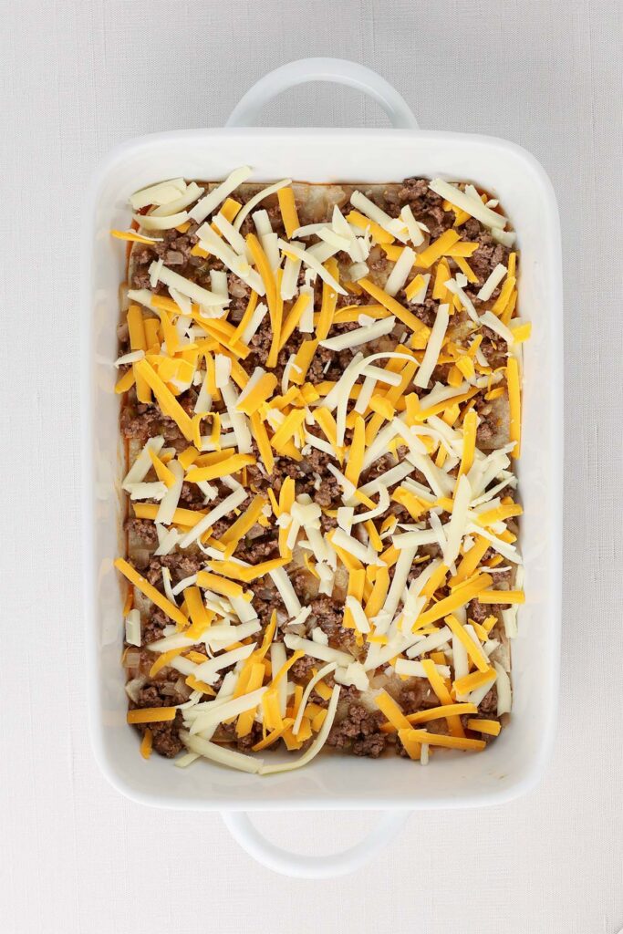 Top down image showing a white baking pan filled with enchilada sauce and a layer of cut up tortillas and ground beef with shredded cheese