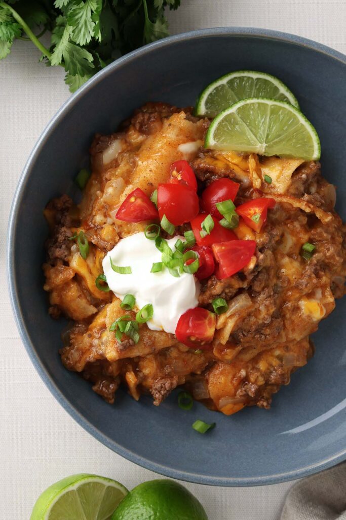 Top down image showing a blue bowl filled with an enchilada dish that is topped with sour cream, green onion, diced tomatoes and lime wedges