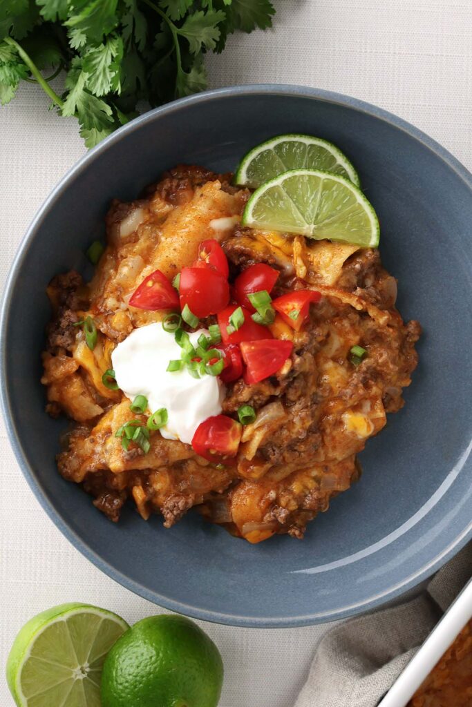 Top down image showing a blue bowl filled with an enchilada dish that is topped with sour cream, green onion, diced tomatoes and lime wedges