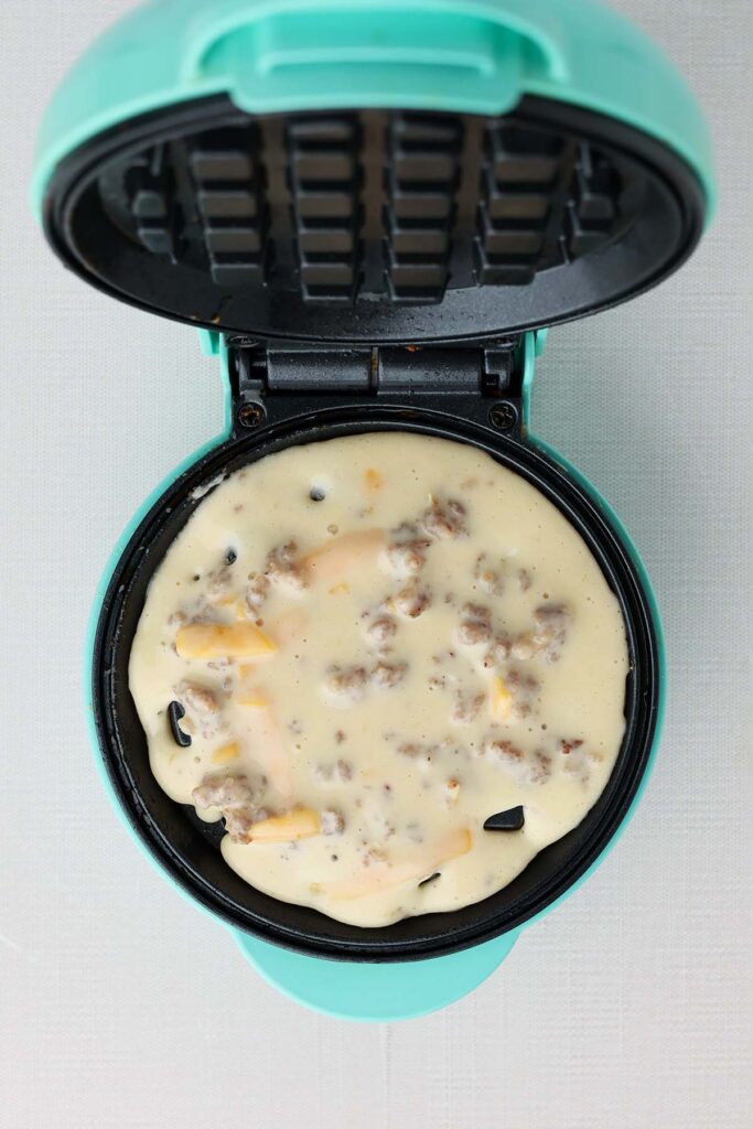 Top down image showing a mini waffle maker filled with batter that is loaded with cheese and sausage