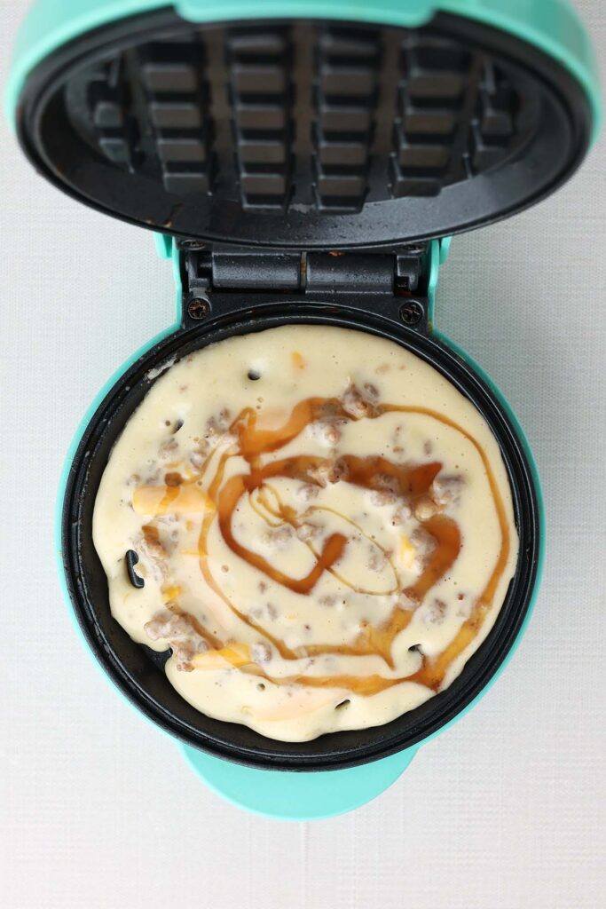 Top down image showing a mini waffle maker filled with batter that is loaded with cheese and sausage with a drizzle of syrup