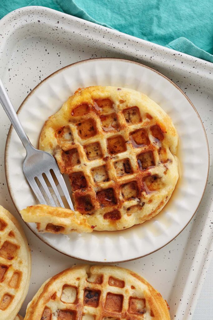 Close up image showing a waffle on a white plate smothered in syrup with a bite on a fork