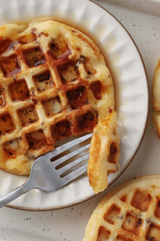 Close up image showing a waffle on a white plate smothered in syrup with a bite on a fork