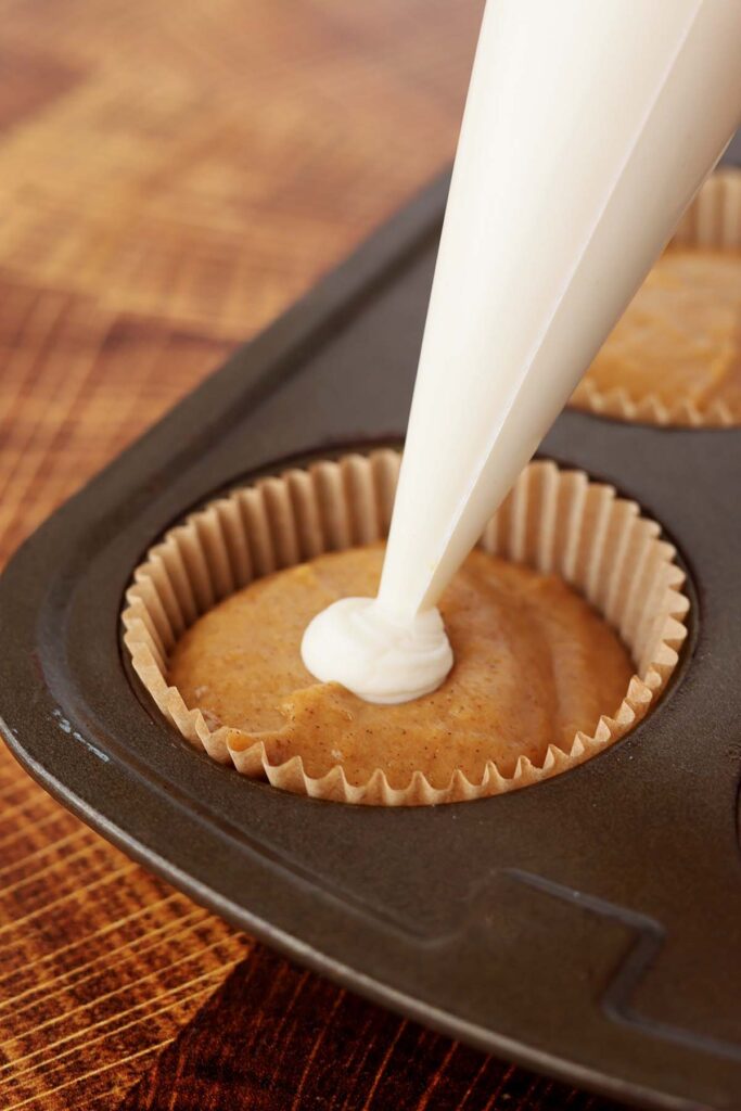 close up showing a cupcake liner inside of a cupcake tin filled with cake batter and a cream cheese mix being pipped in