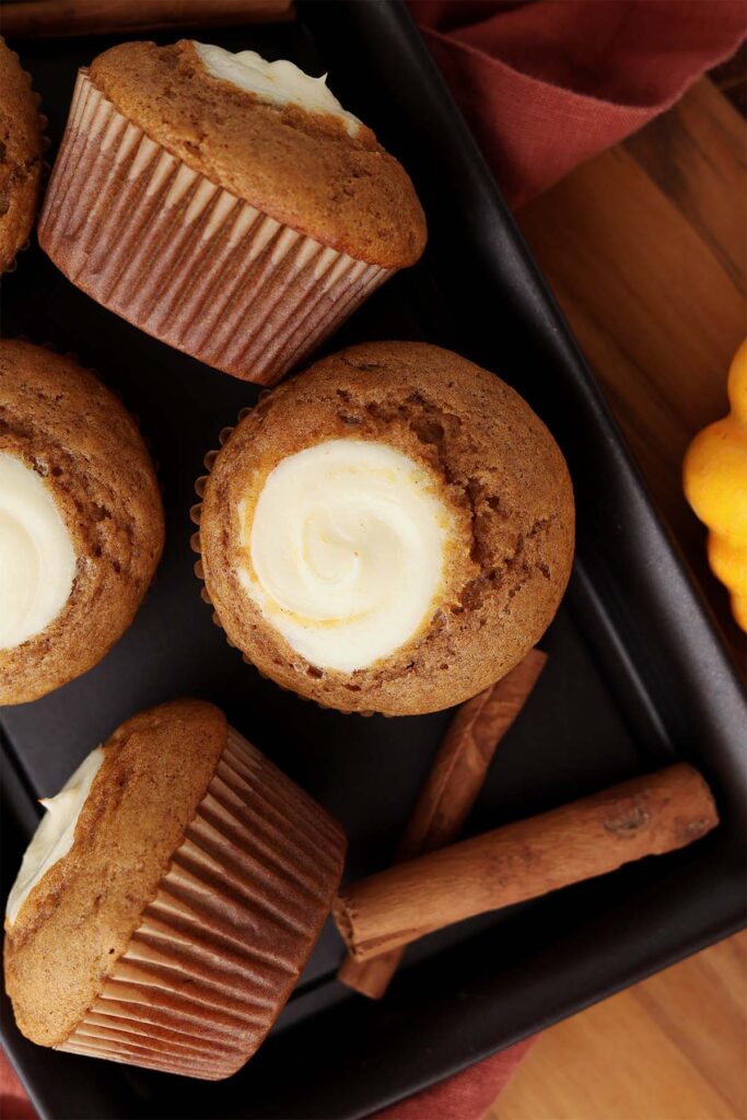 Top down image showing a spread of cupcakes that have a swirl of cream cheese in the middle on a black tray with cinnamon sticks and a rust colored napkin off to the side