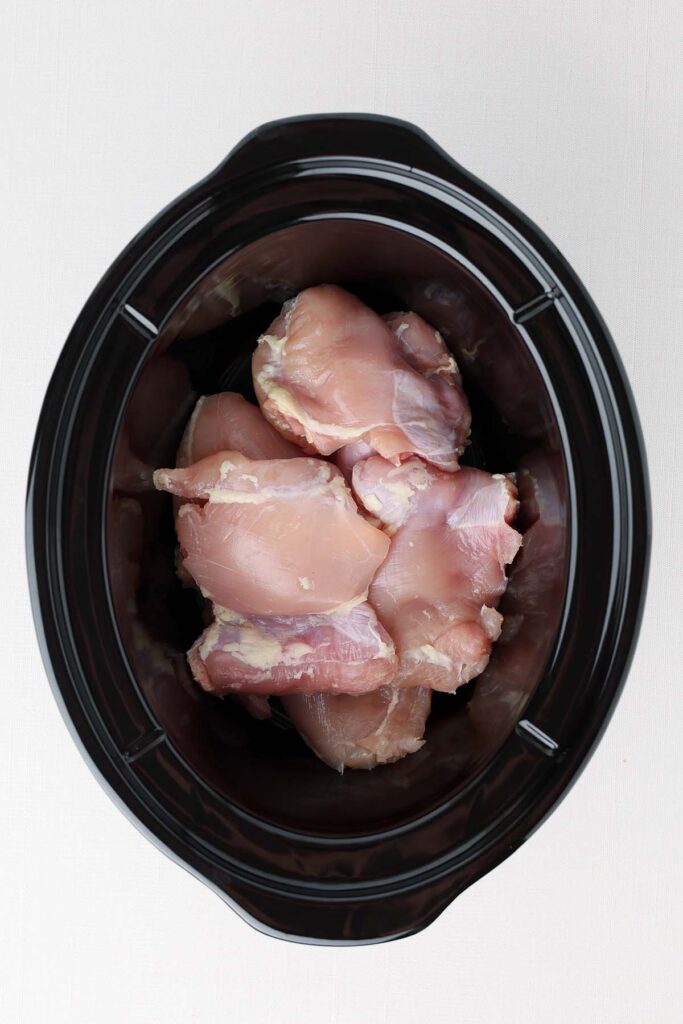 Top down image showing the inside of a black slow cooker piled with chicken thighs