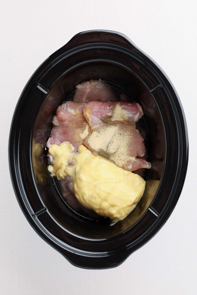 Top down image showing the inside of a black slow cooker with chicken thighs, a can of cream of chicken soup, and seasonings