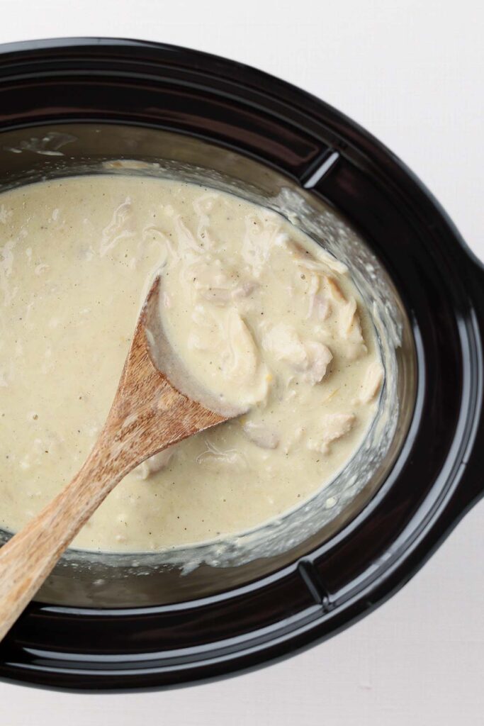 Top down image showing the inside of a black slow cooker with a creamy gravy and chicken pieces with a wooden spoon