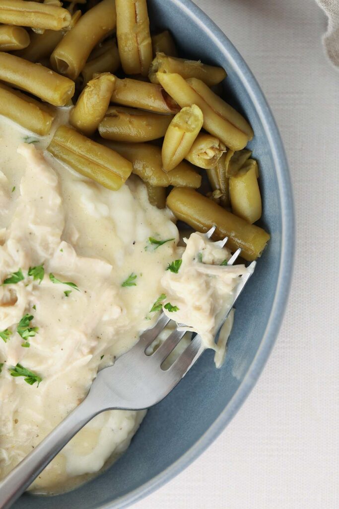 Top down image showing a blue shallow bowl filled with mashed potatoes and chicken gravy with green beans a bite on a fork